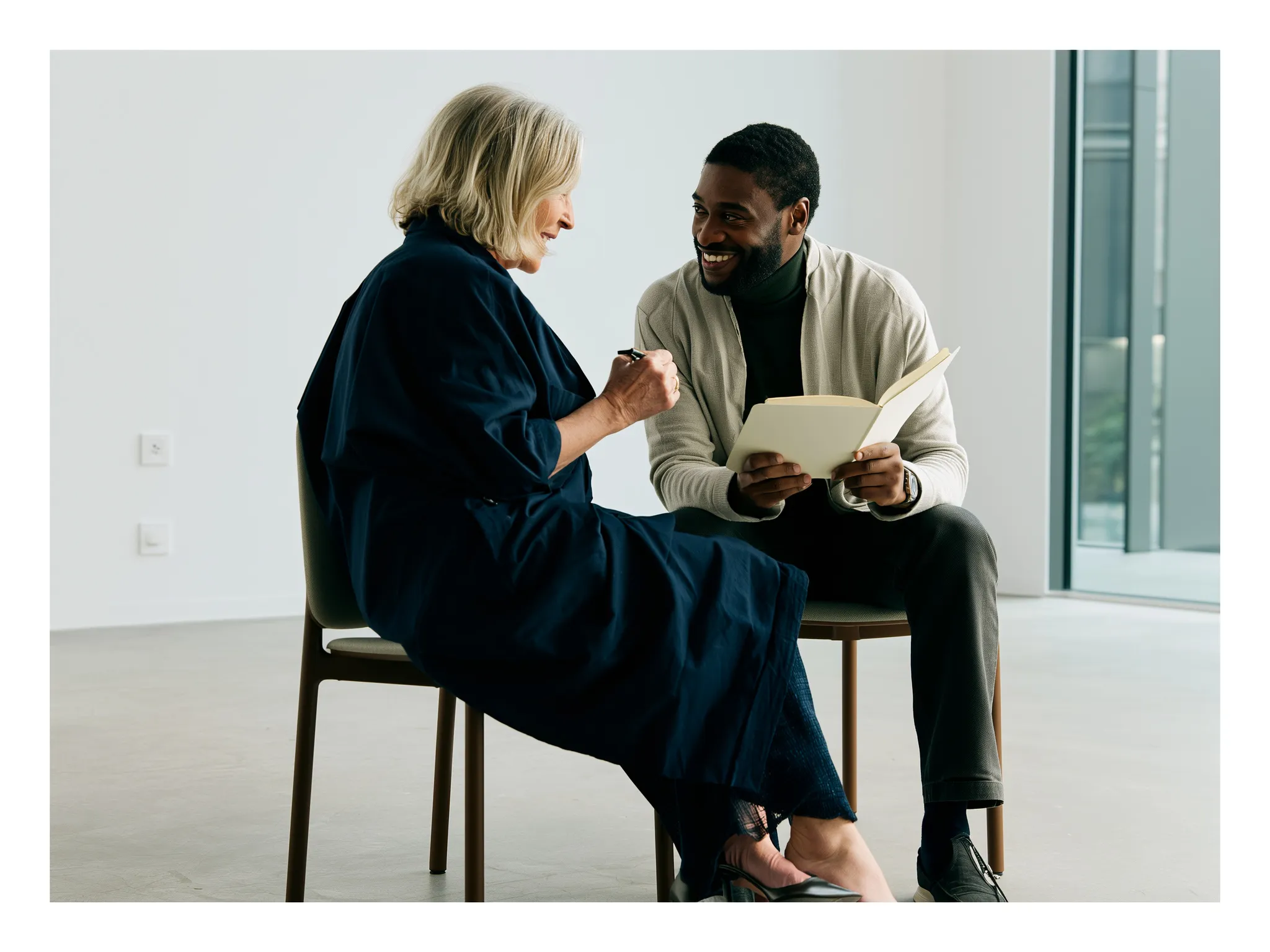 A poet writing in a notebook while listening closely to a participant in a bright room